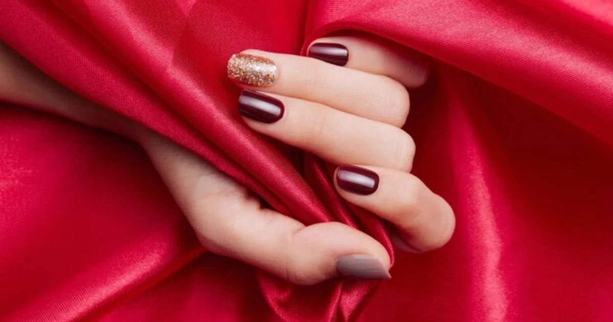 A hand with elegant Valentine's Day nails: burgundy polish with gold glitter accent on a red background. 43 Red Nail Ideas For a Chic Bold Manicure