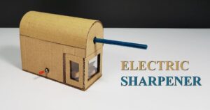 A homemade cardboard model of an electric pencil sharpener with a blue pencil inserted. It has a visible window for the sharpening mechanism and a red toggle switch on the side. The words "ELECTRIC SHARPENER" are on the right. How Does an Electric Pencil Sharpener Work_ in 5 Simple Steps