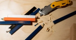 A close-up shows several carpenter pencils in orange, white, black, and blue on a wooden surface. A yellow and grey utility knife is near a partially sharpened blue pencil, with wood shavings scattered around.