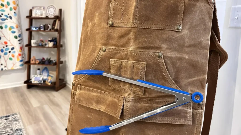Close-up of a brown woodworking apron with blue-tipped metal tongs attached by a strong magnet.