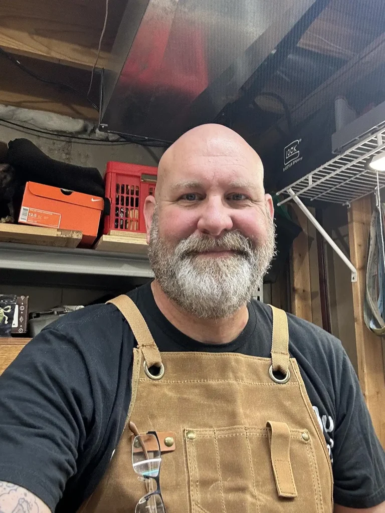 Selfie of a smiling person with a beard wearing a brown woodworking apron in a workshop.